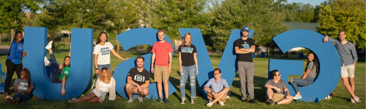 Students pose with the bright blue "GVSU" letters in front of the Cook Carillon Clock Tower on Grand Valley State Unviersity's Allendale Campus.